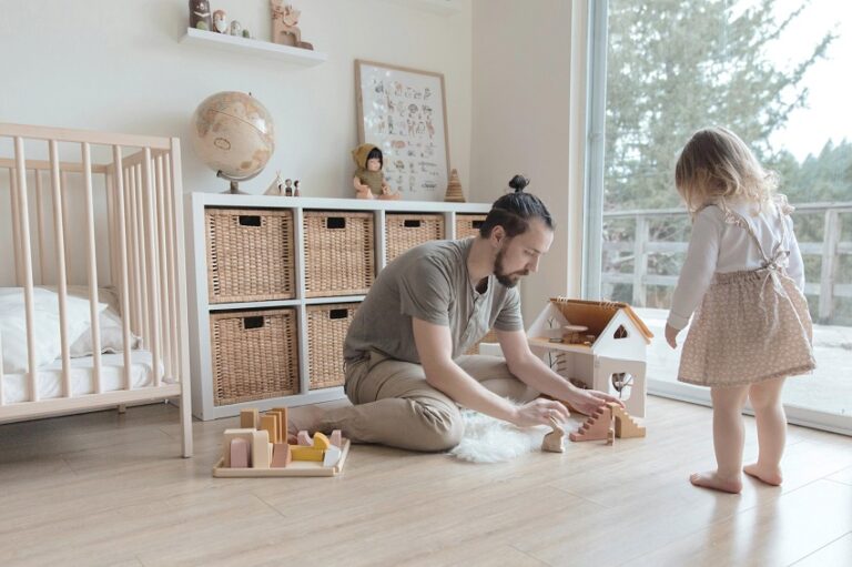 picture of a man kneeling on the floor he is playing by building a toy house while his toddler duaghter observes him