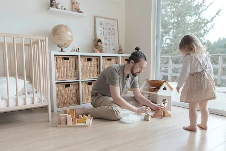 picture of a man kneeling on the floor he is playing by building a toy house while his toddler duaghter observes him