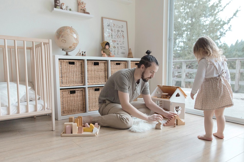 picture of a man kneeling on the floor he is playing by building a toy house while his toddler duaghter observes him